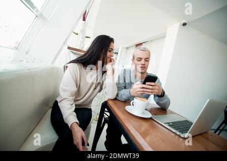 Young couple meets in coffeehouse. Blonde man showing something fun in his smartphone to brunette attractive woman. Modern laptop on table. Social med Stock Photo