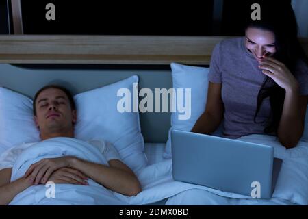 Woman sitting on bed in the night and using laptop computer while husband sleeping beside her. Stock Photo