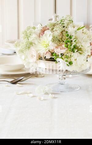 Floral decorations on summer party table. Candle in glass vase, flower wreath around it. White plates, table cloth, romantic wedding reception. Stock Photo