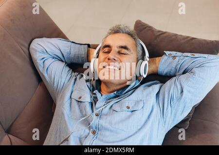 Middle aged man listening to music, lying on sofa with closed eyes and smiling with teeth. Casual style. Man enjoying his favorite music and relaxing Stock Photo