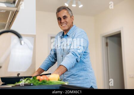 Middle aged man enjoys cooking in his modern kitchen. He stands near stove, waits for the dish to be ready, looks at camera and smiles. Blurred colour Stock Photo
