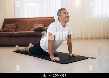 Senior man meditating while doing cobra pose in living room. Stretching exercises. Indoor shot of happy man practicing yoga. Morning workout. Plastic Stock Photo
