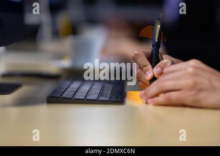 Close up image of businessman writing on an adhesive note at table in office at night, focus on hand. Stock Photo