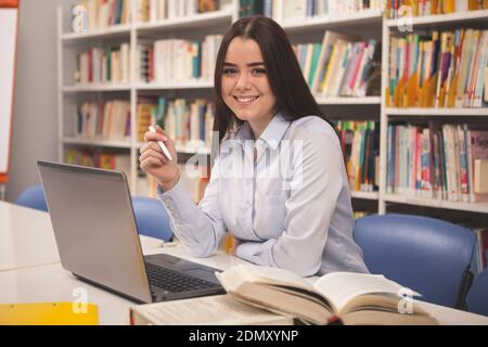 Happy beautiful student girl smiling to the camera, while studying at the library. Gorgeous female students working on her laptop at the college libra Stock Photo