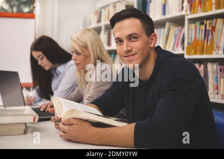 Handsome young man smiling to the camera, while studying with his classmates at the college library. Cheerful male student reading a book, preparing f Stock Photo