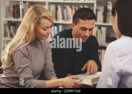 Beautiful blond haired female student studying with her classmates, reading a book together at the library. Group of college friends enjoying working Stock Photo