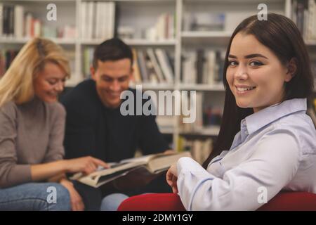 Lovely happy female student smiling to the camera, sitting relaxed at the college library, her classmates reading on the background. Cheerful woman re Stock Photo