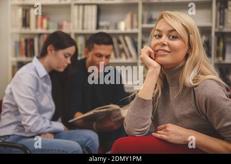 Gorgeous happy blond haired woman smiling to the camera, relaxing at college library with her friends tudying on the background. Lovely female student Stock Photo