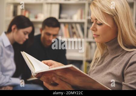 Cropped close up of a beautiful female student reading a book attentively, her college friends studying on the background. Charming woman enjoying rea Stock Photo