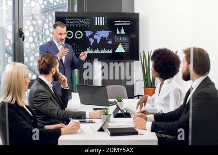 Serious mature businessman manager stands near big digital wall screen with company financial and growth charts and talks to his multiethnic Stock Photo