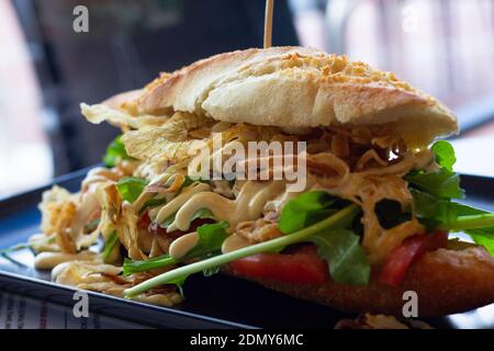Enormous French baguette bread chicken sandwich with vegetables, fried onion and mayonnaise on black plate. Delicious healthy meal, gourmet bar Stock Photo