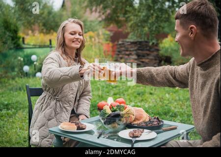 A young couple celebrates a family holiday, Dating and wedding anniversary. A man and a woman clink glasses of beer while sitting at a table during a barbecue on a cool autumn day Stock Photo