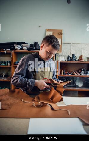 In a boot shop, an experienced young master cuts out leather shoe elements with scissors. Stock Photo