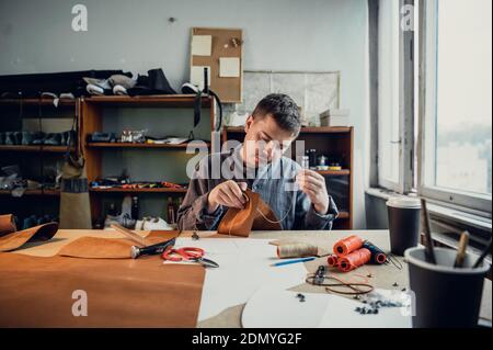 Sitting at a table in the workshop a young guy manually sews leather elements to each other. Stock Photo