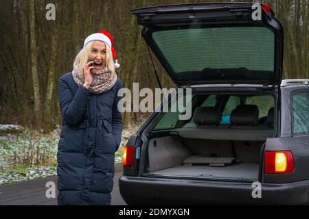 Young blonde woman in a Santa Claus hat is talking on a mobile phone in front of a car with the trunk up and the lights on and shouts into the phone Stock Photo