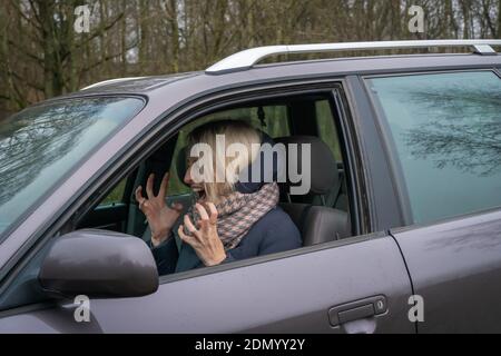 Young blonde woman in a down jacket and a voluminous scarf sits at the wheel of a car and shouts while gesturing with her hands. Stock Photo