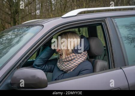 Young blonde woman in a down jacket and a voluminous scarf sits at the wheel of a car and shouts while gesturing with her hands. Stock Photo