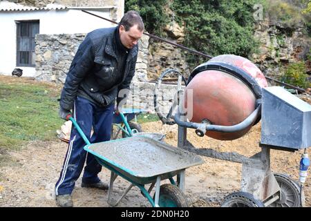 Bricklayer next to concrete mixer carrying a concrete wheelbarrow. Stock Photo