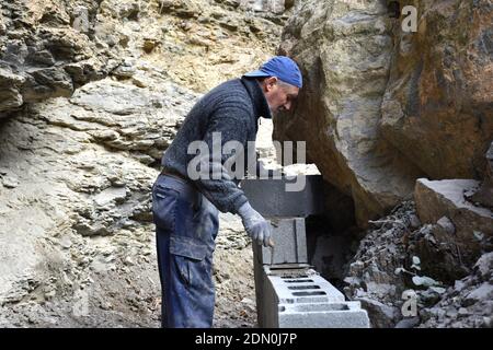 Elderly worker building a wall of blocks and concrete next to large stone rock. Stock Photo