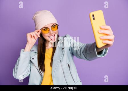 Photo of stylish pretty youngster lady hold telephone smiling sticking tongue make selfies followers blogger wear sun specs casual hat jacket yellow Stock Photo