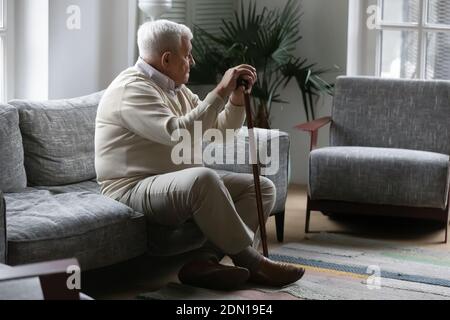 Thoughtful mature male resting on sofa leaning on walking stick Stock Photo