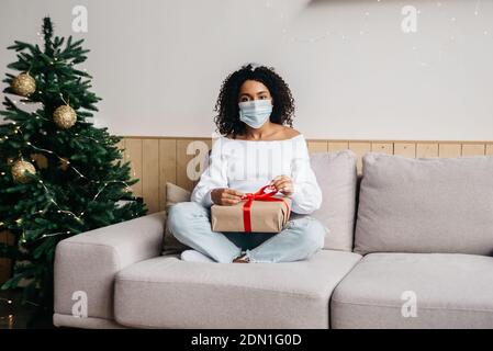 Millennial african american woman wearing a protective mask ah holdig box with red ribbon. Health protection during Coronavirus Christmas celebrating Stock Photo