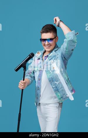 handsome vocalist man in denim and sunglasses singing to microphone on blue background. Stock Photo