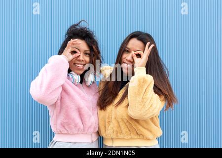 Portrait of two smiling friends putting their fingers close to their faces forming the symbol of okay. They are on a blue background. Space for text. Stock Photo