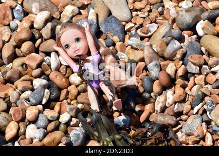 Small discarded doll lying on colorful pebble beach with seaweed, Brighton, Sussex, England, United Kingdom Stock Photo