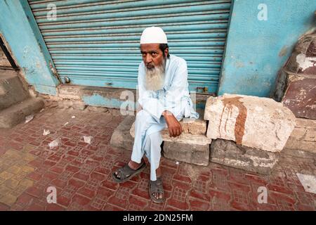 Jamnagar, Gujarat, India - December 2018: A portrait of an elderly Indian man with a beard wearing a white skull cap sitting on the pavement. Stock Photo