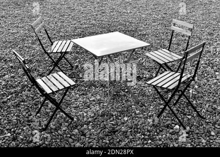 Seating arrangement with four empty chairs around a square table on pebbled beach after the rain in black and white, Brighton, Sussex, England, UK Stock Photo