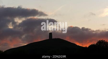 the sun setting behind Glastonbury Tor, Somerset, England, UK, on a summers evening and lighting up the clouds Stock Photo