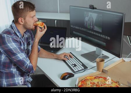 Young man looking concentrated, working on his computer and eating pizza. Young male freelancer eating fastfood in front of computer Stock Photo