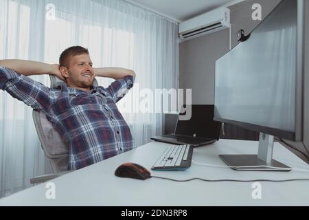 CHeerful relaxed program developer stretching his back, sitting in front of the computer, copy space. Happy male student relaxing after finishing his Stock Photo