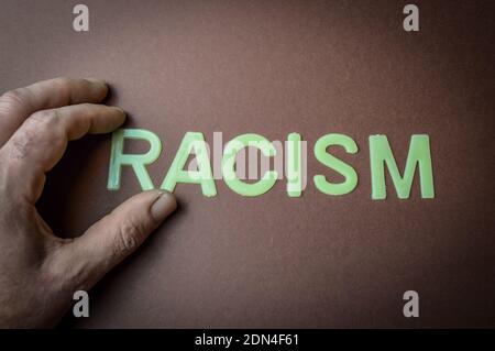 Human fingers holding the word Racism written with plastic letters on a brown paper background, concept Stock Photo