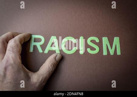 Human fingers holding the word Racism written with plastic letters on a brown paper background, concept Stock Photo