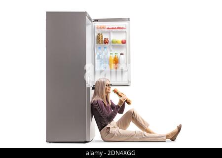 Woman sitting on the floor, leaning on a fridge and eating a sandwich isolated on white background Stock Photo
