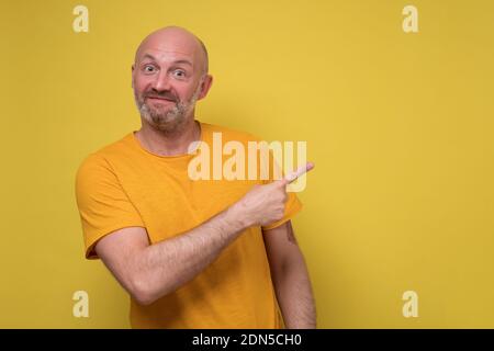 Positive mature woman looking at camera, smiling and pointing aside with hand. Studio shot on yellow wall. Stock Photo