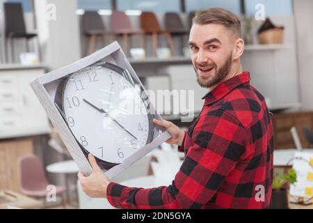 Handsome bearded man looking excited, holding big wall clock. Attractive young man shopping for home goods and decor Stock Photo