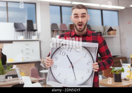 Handsome bearded man looking overwhelmed, holding big wall clock at the furniture store, copy space. Attractive man looking shocked, holding big clock Stock Photo
