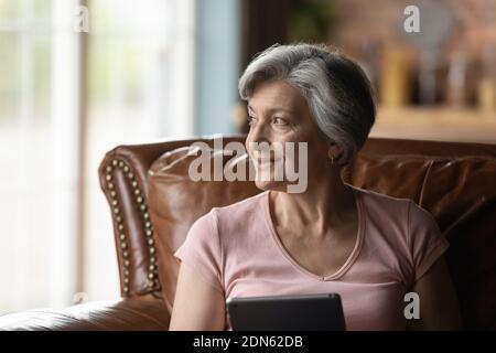 Happy young dreamy older 60s senior woman using tablet app. Stock Photo
