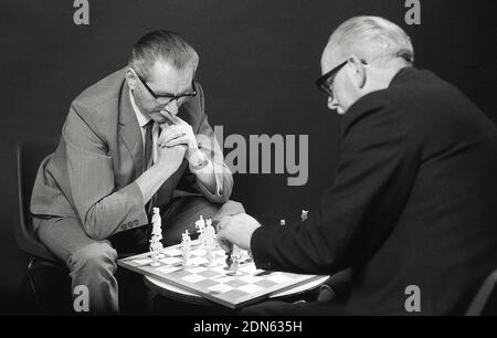 1960s, historical, two older men concentrating, sitting opposite each other playing a game of chess, with the chessboard sat on a small coffee table, between them, England, UK. A popular board game, an abstract strategy game, the modern version dating back to the 15th century, chess is played on a 64 square chessboard with 16 pieces, the winner attacks and checkmates the opponent's king, so there is no way on the board for it to escape. As the game involves a combination of logic, patterns, focus, memory and decision making, playing chess is good exercise for the brain. Stock Photo