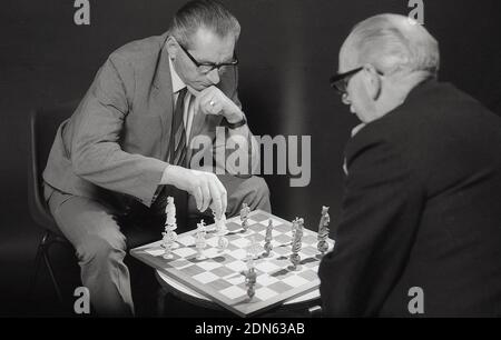1960s, historical, two older men sitting opposite each other playing a game of chess, with the chessboard sat on a small coffee table, beween them, England, UK. A popular board game, an abstract strategy game, the modern version dating back to the 15th century, chess is played on a 64 square chessboard with 16 pieces, the winner attacks and checkmates the opponent's king, so there is no way on the board for it to escape. As the game involves a combination of logic, patterns, focus,memory and decision making, it is said that playing chess is good exercise for the brain. Stock Photo