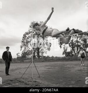 1965, outside in a sports field, watched by a fellow schoolboy, a young man doing the high jump at a high school sports day, using a form of the straddle method, the parallel straddle, where the lead leg is kicked high in the air and the head and trunk clear the bar at the same time. The 1956 Olympic Champion, Charles Dumas - and the first jumper to clear 7ft - used this version of the straddle. The other version of the straddle is the dive method, where the head goes over the bar first. In amateur high jumping the two styles are often mixed together, in a what ever gets you over bar works. Stock Photo