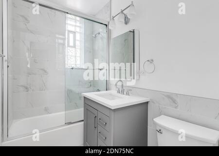 A bright, white bathroom with a grey cabinet, marble tiled shower and a glass sliding door. Stock Photo