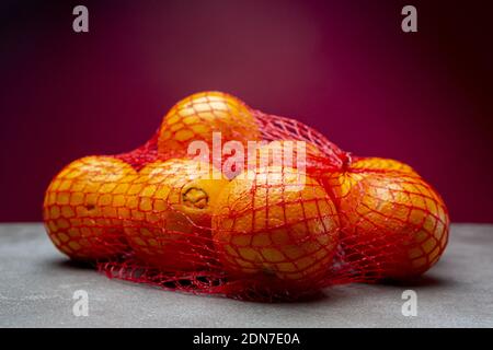 Bunch of oranges in red carrying net on a textured kitchen surface and dark purple background Stock Photo