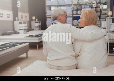 Rear view shot of a senior couple hugging, smiling at each other sitting on a bed at furniture store. Loving happy elderly husband and wife embracing, Stock Photo