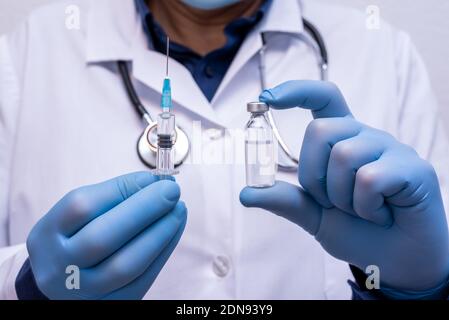 A physician wearing blue gloves holds a syringe ready for injection in one hand and in the other hand holds a container containing a vaccine Stock Photo
