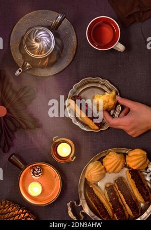 Tasty eclairs or brewing cakes and pewter tea pot and cup of tea. Natural Christmas decor, candles, red berries. Christmas top view on dark linen Stock Photo