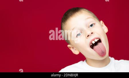 Boy teenager 7-10 in a white t-shirt makes faces, showing tongue, on a red background. Banner Stock Photo